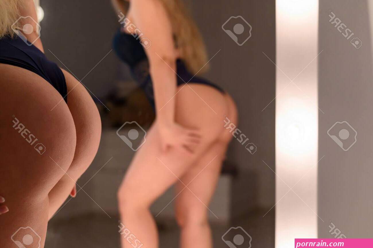 A Blonde With Long Hair In Black Underwear Is Standing By The Mirror. Rear View Of A Girl In Thong. Reflection Of A Woman With Wide Hips With A Big Booty And